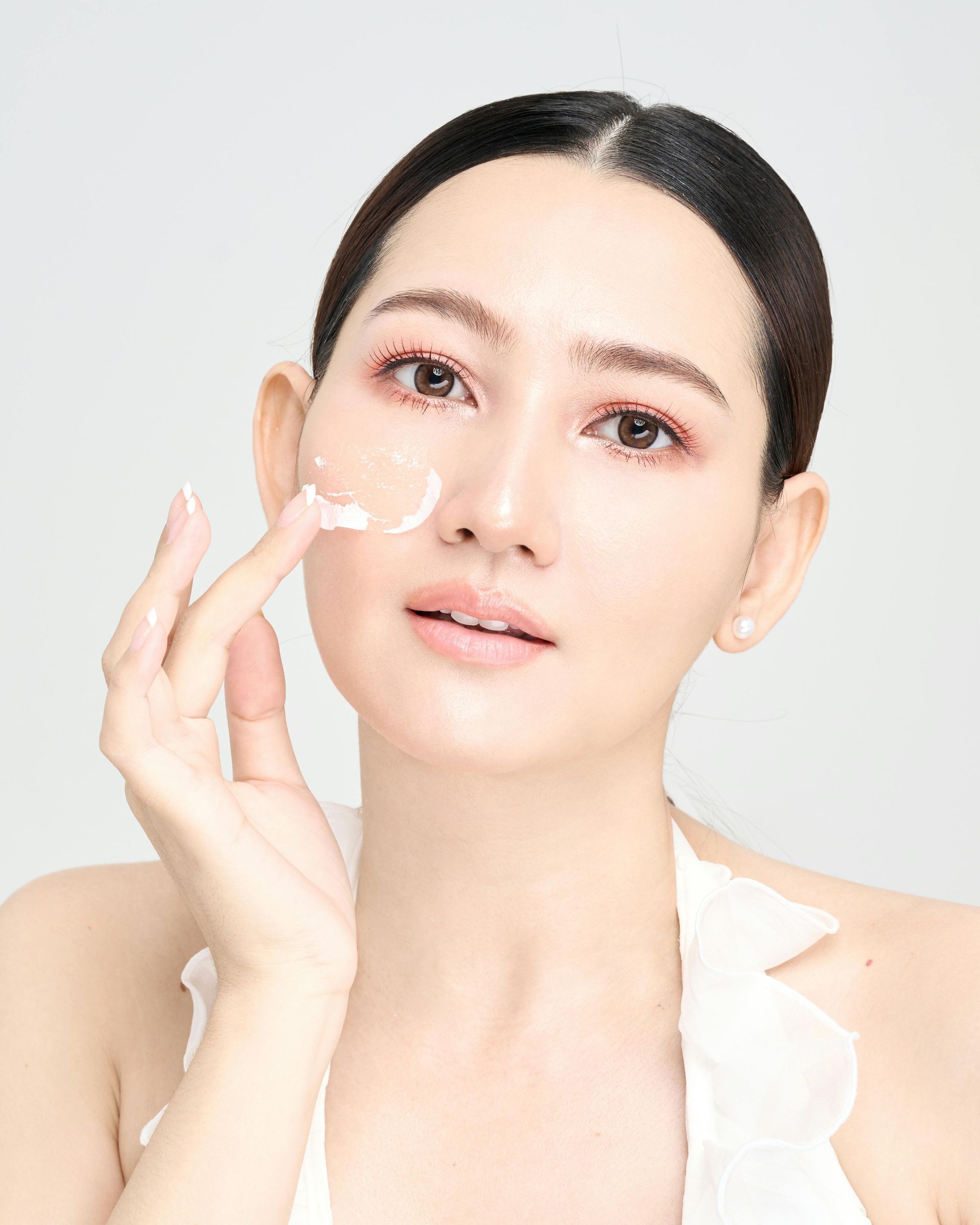 Woman applying cream to her face with a finger, looking at the camera. She has fair skin and is wearing a white top.