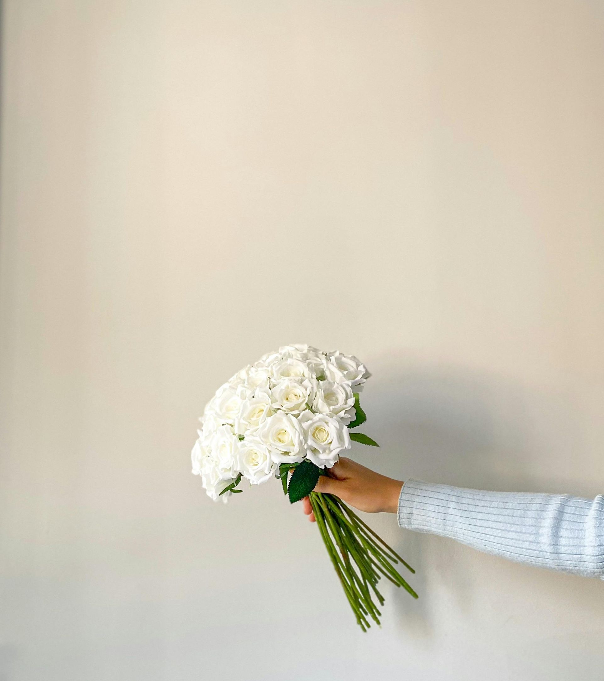 Person holding a bouquet of white flowers against a cream-colored wall. The person wears a light blue sweater.