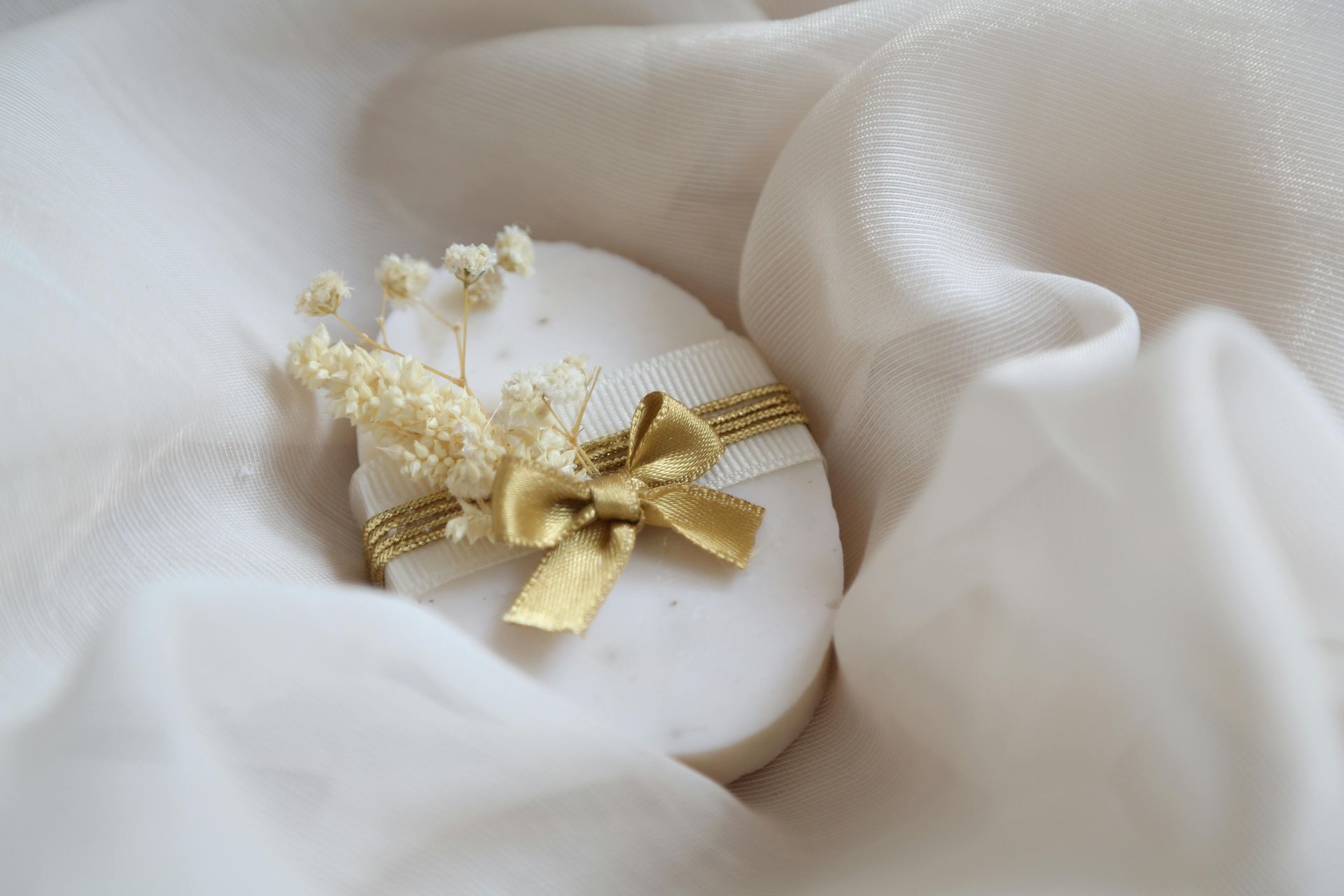 Oval, white soap decorated with a gold ribbon and dried flowers, resting on white silk.