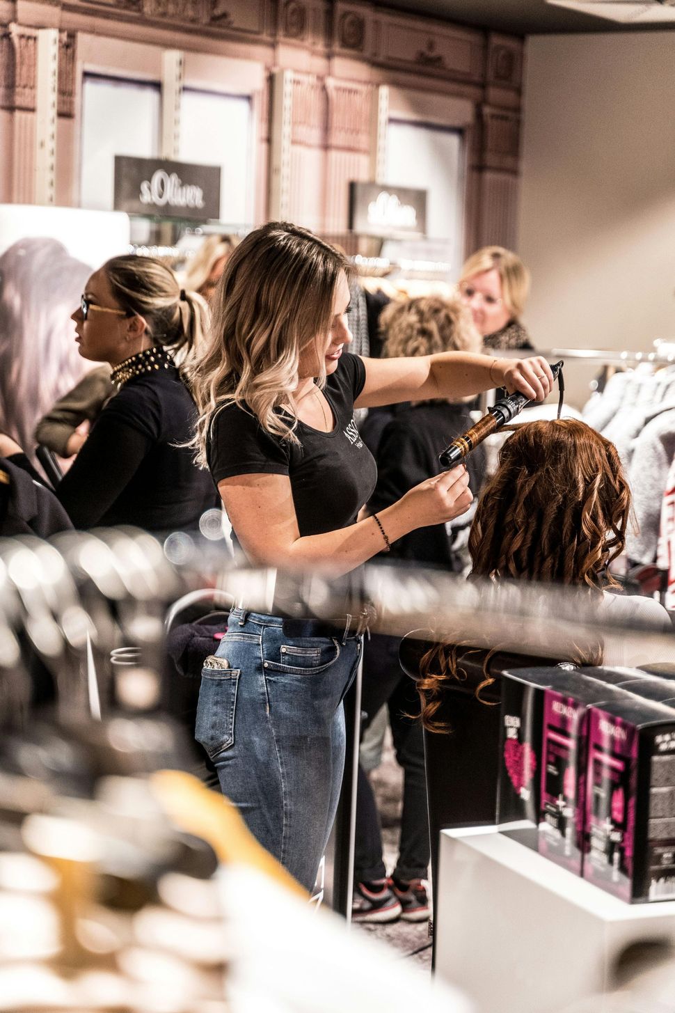 A woman with light brown hair using a curling iron on a mannequin head in a clothing store. Others are in the background.