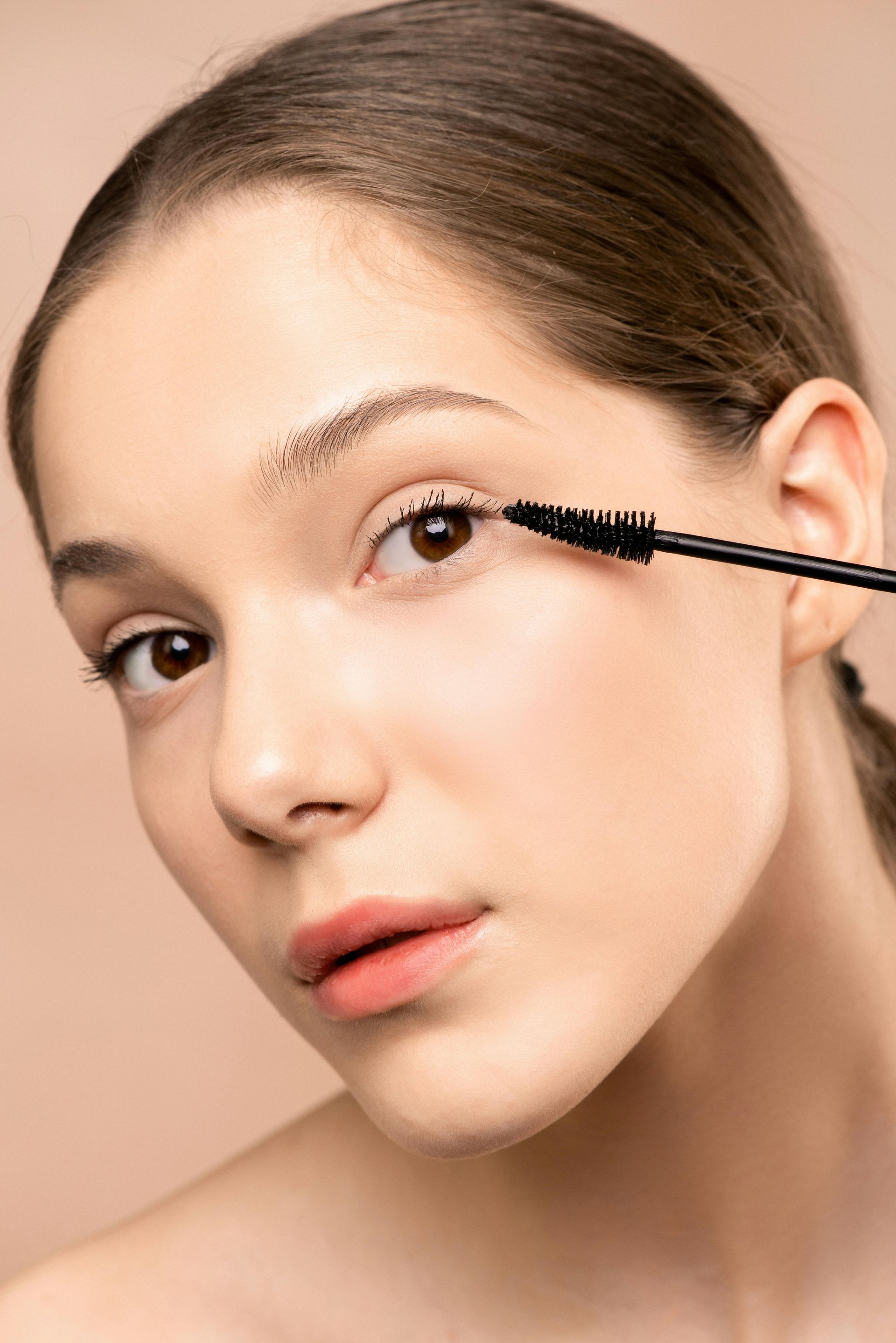 A woman applies mascara to her eyelashes. She looks towards the camera with a neutral expression; beige background.