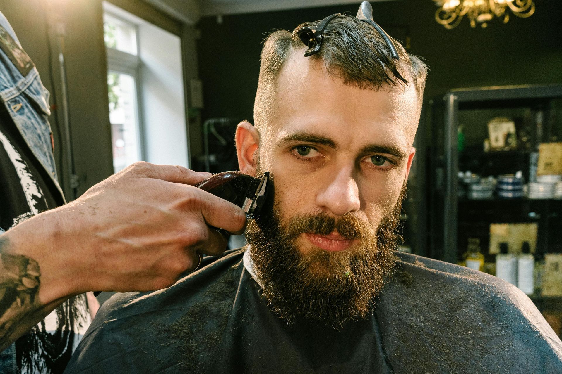 A man with a beard getting his sideburns trimmed with clippers at a barbershop.