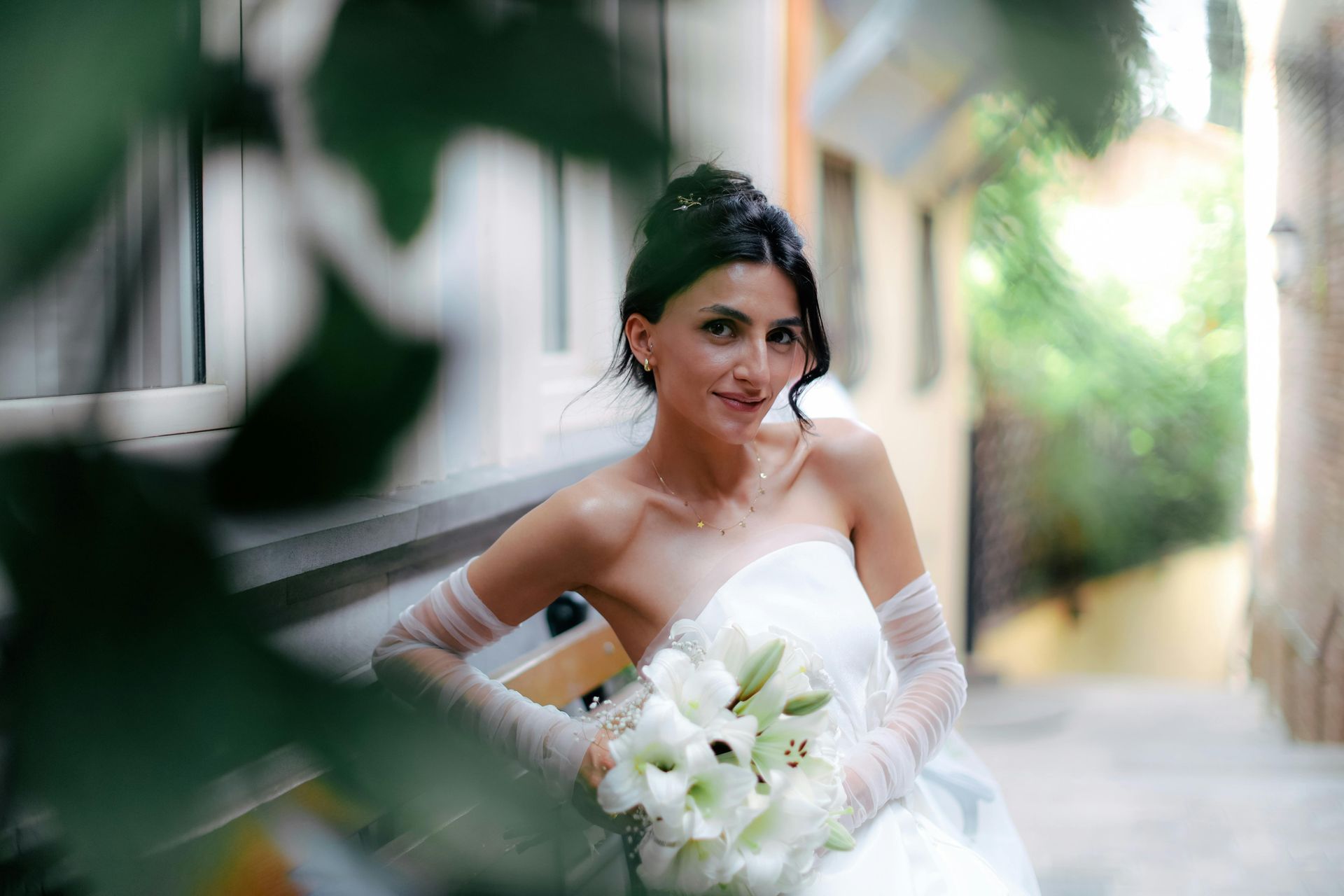 Bride in a strapless white dress, holding lilies. She smiles gently, posing outdoors in an alley.
