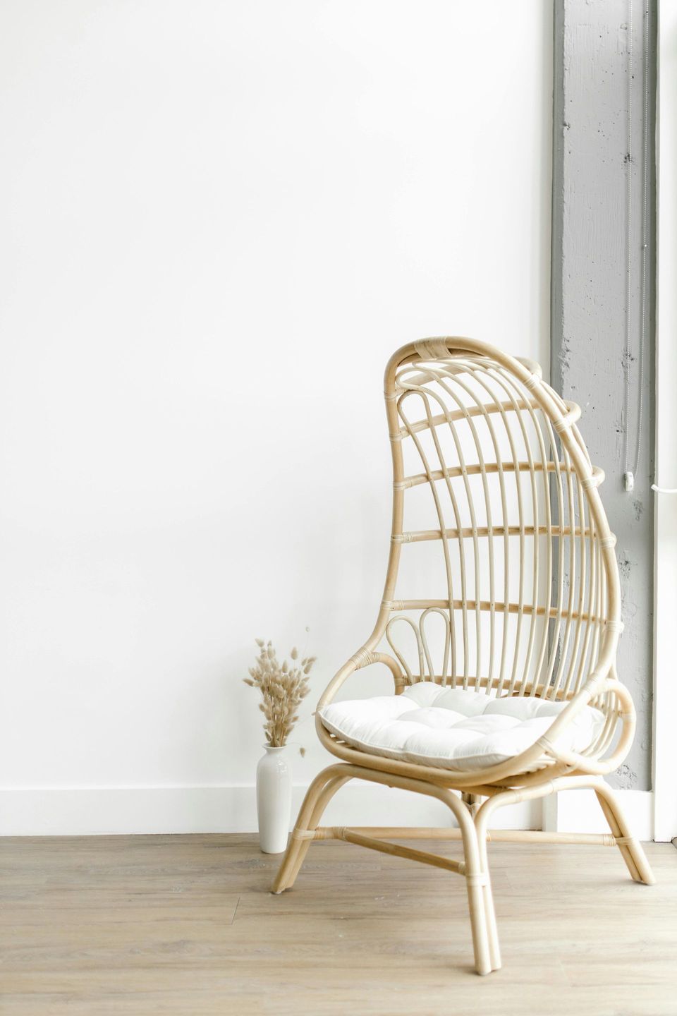 Wicker teardrop chair with white cushion sits against a white wall, near a vase with dried flowers.