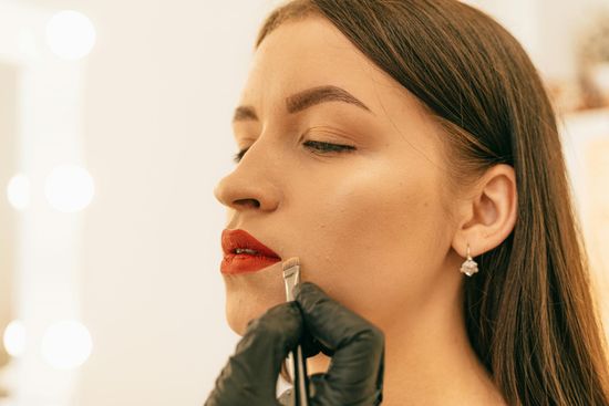 A woman's profile as a gloved hand uses a tool to apply lip filler. She has red lipstick, and is in a bright setting.