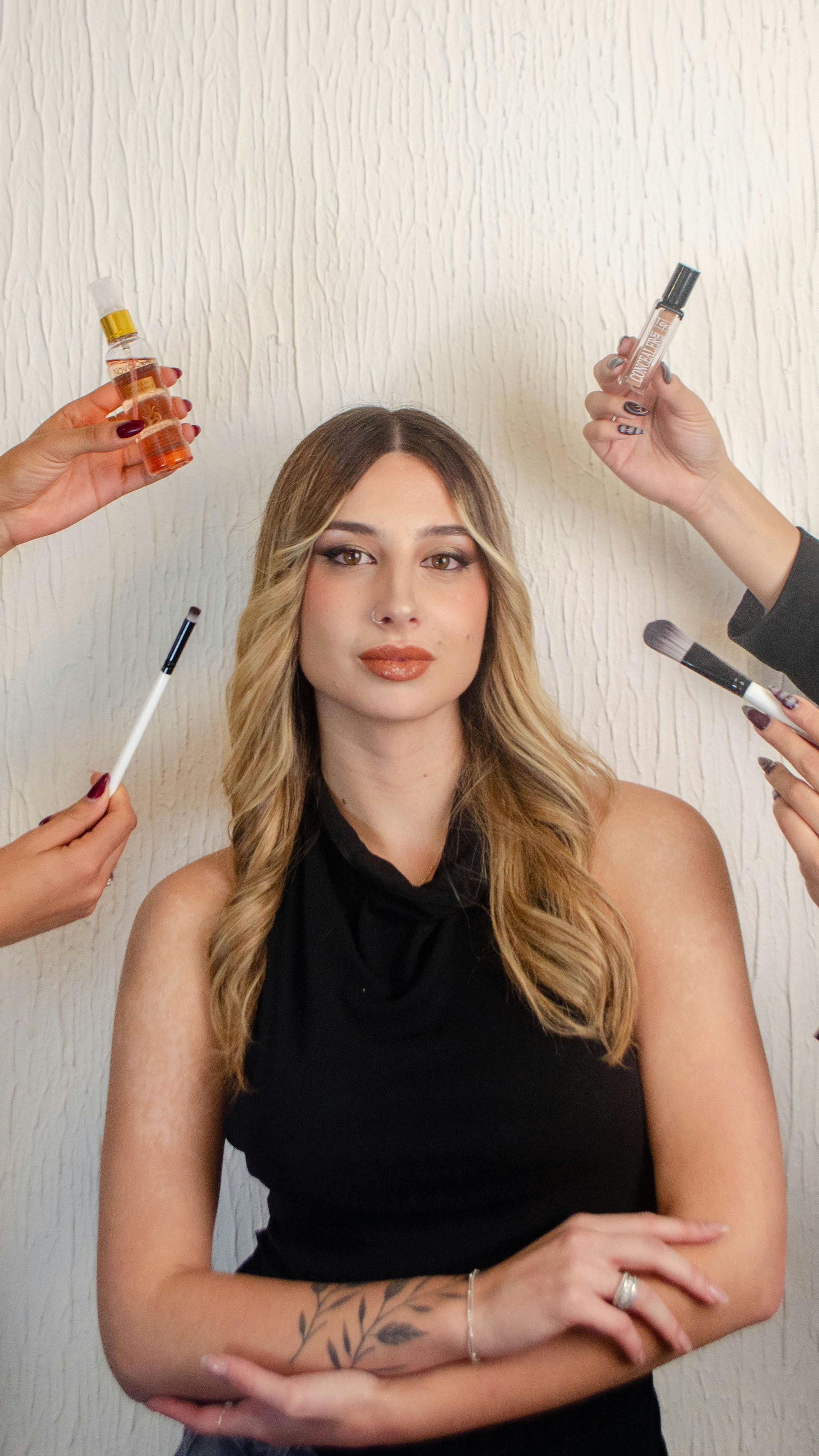 Woman with long blonde hair and a black top, surrounded by hands holding makeup products.