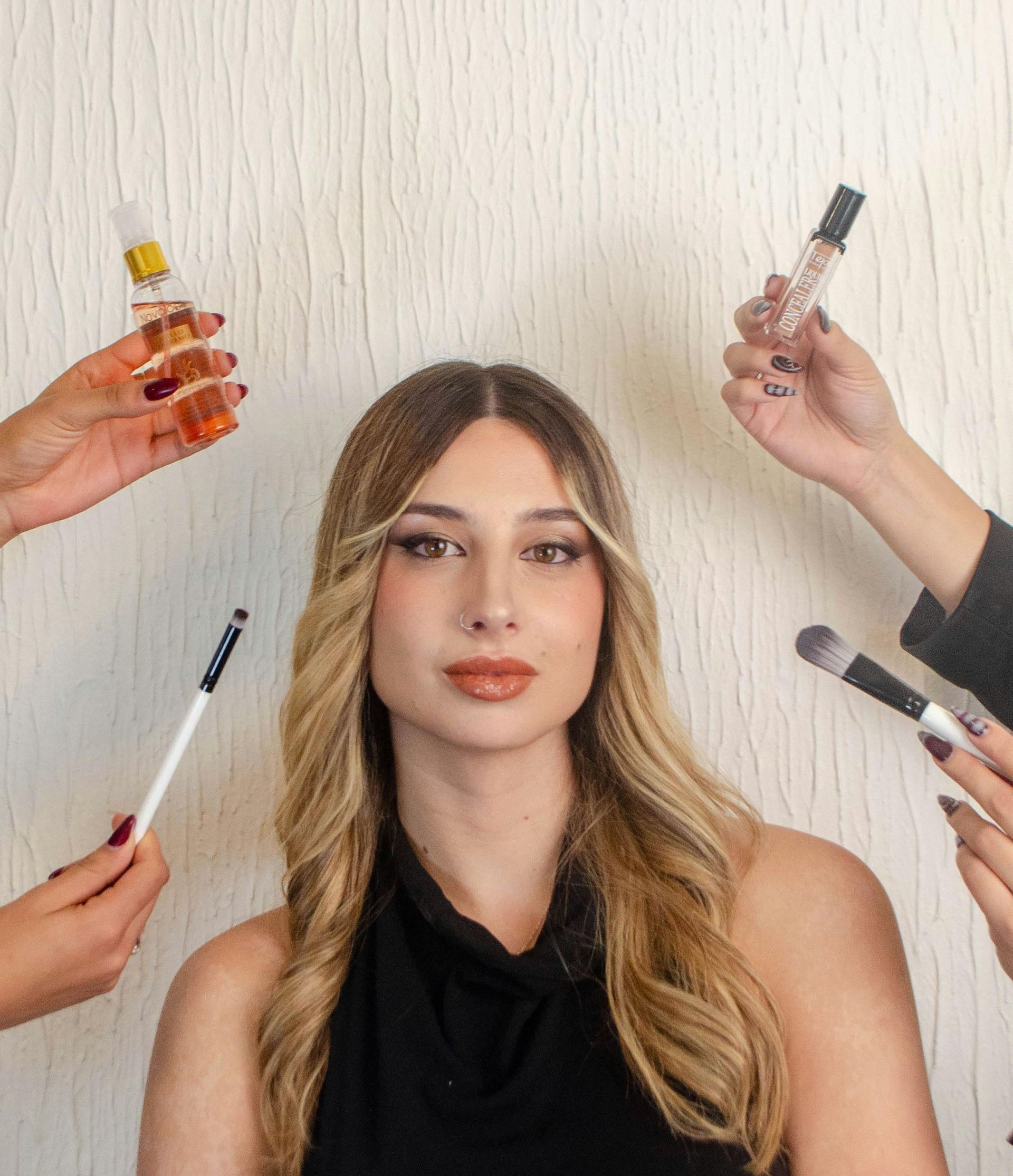 Woman with blonde hair and makeup, surrounded by hands holding beauty products against a neutral background.
