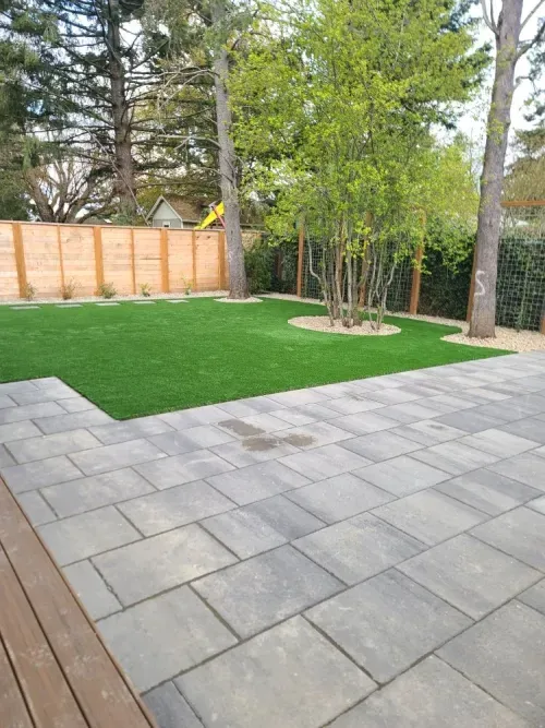 A backyard with a patio , grass , trees and a wooden fence.