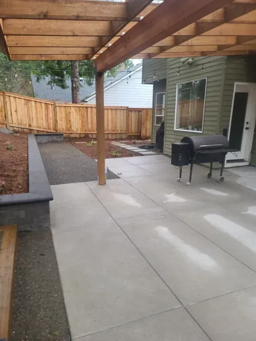 A patio with a grill under a pergola in the backyard of a house.