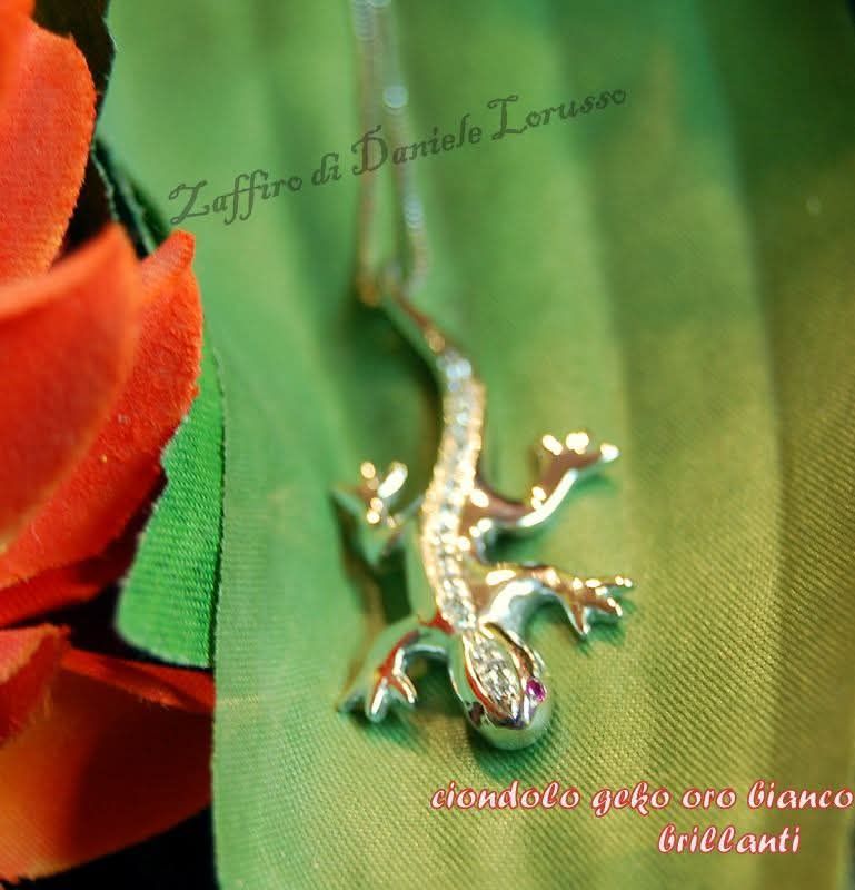 Ciondolo a forma di geco in argento con pietre preziose bianche su sfondo verde e arancione.