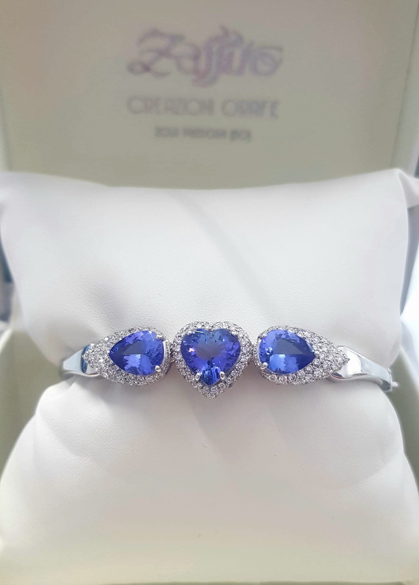 Bracciale con tre cuori di pietre preziose blu, circondati da piccoli diamanti, su un cuscino bianco in una scatola.