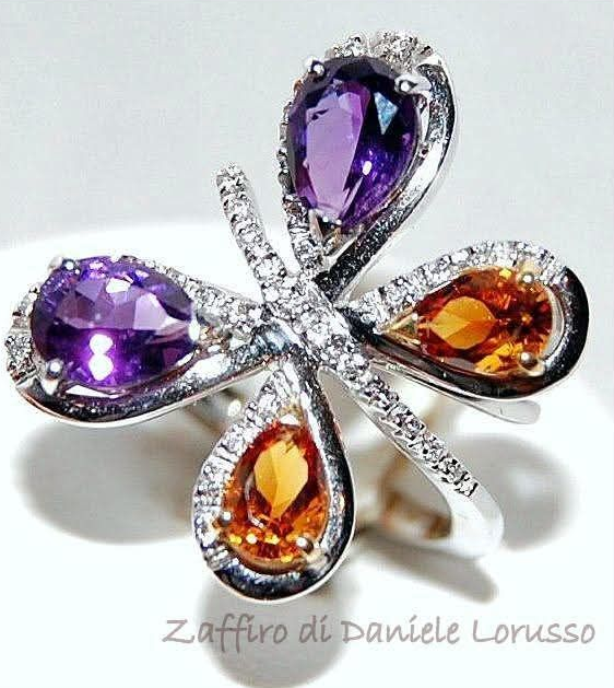 Anello a forma di farfalla con gemme di ametista viola e citrino arancione, circondato da diamanti, su sfondo bianco.