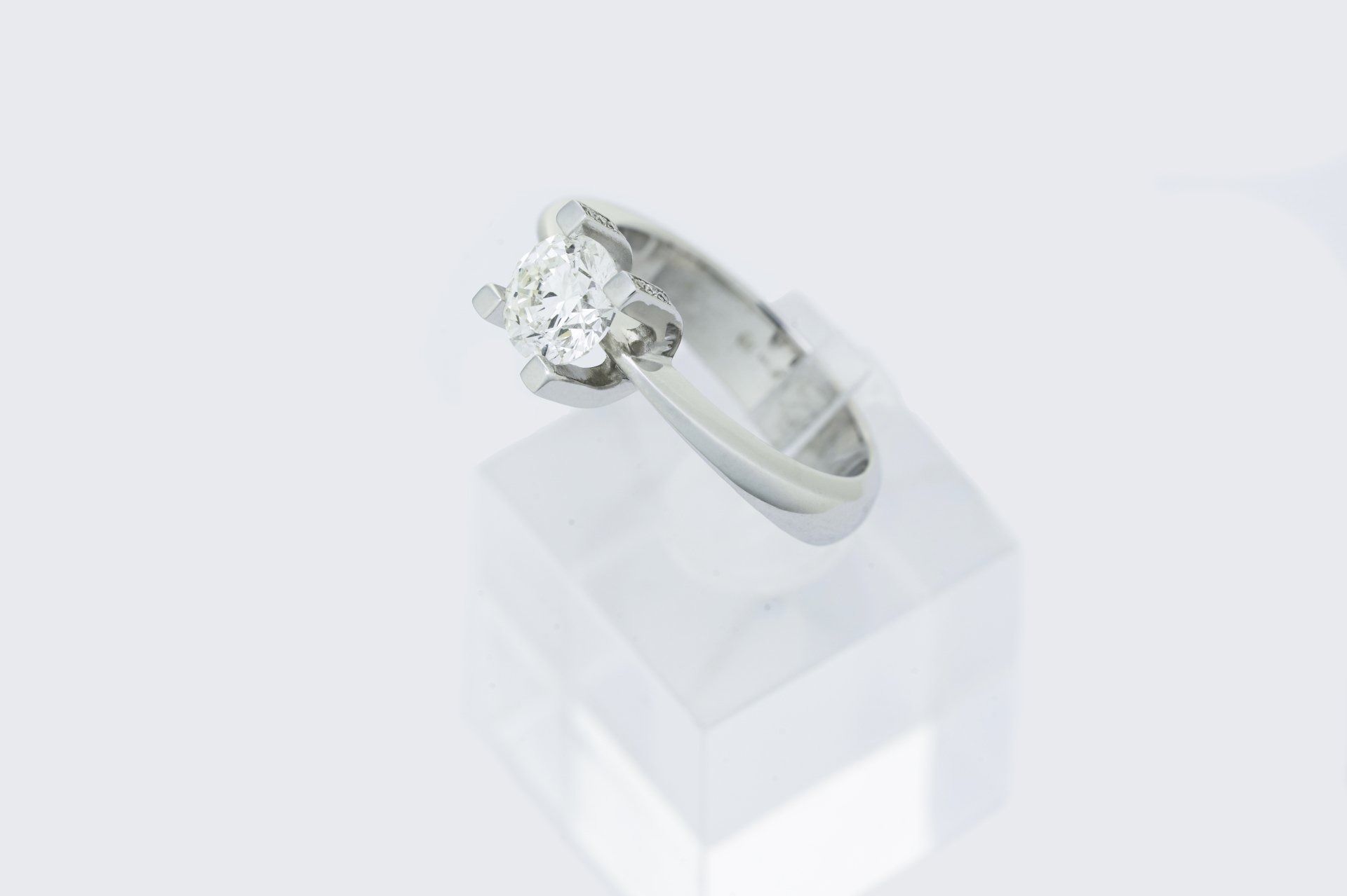 Anello con diamante solitario su supporto trasparente, incastonato su sfondo bianco.