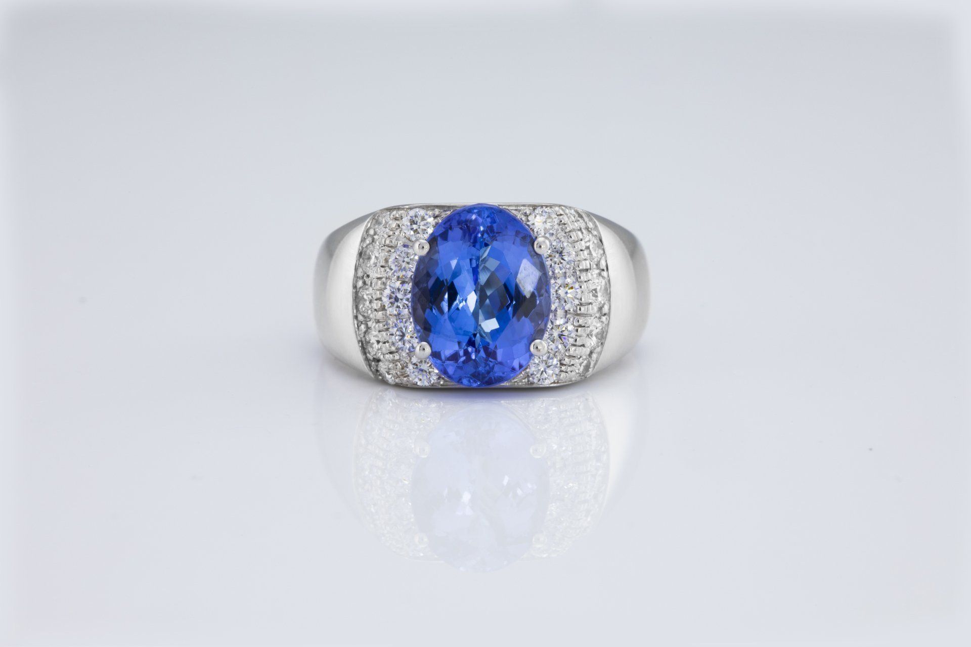 Anello con pietra preziosa ovale blu, circondata da diamanti più piccoli, su una superficie bianca riflettente.