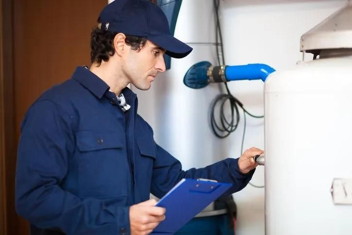 Plumber in blue uniform inspecting a water heater, holding a clipboard in a utility room.