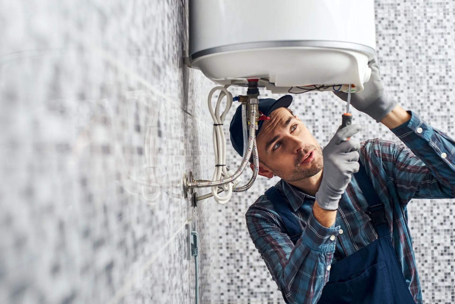 Plumber in blue overalls and cap working on a water heater in a bathroom.