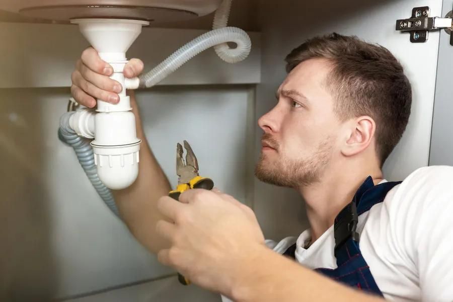 Man in a blue apron fixing sink plumbing with pliers; focused expression.