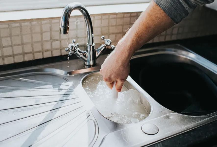 Hand washing a white dish in a stainless steel kitchen sink with running water.