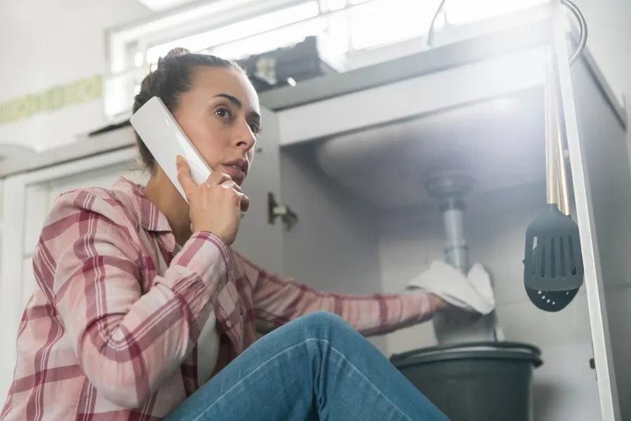 Woman on phone, leaning under sink, likely dealing with a plumbing issue, holding a rag and bucket.