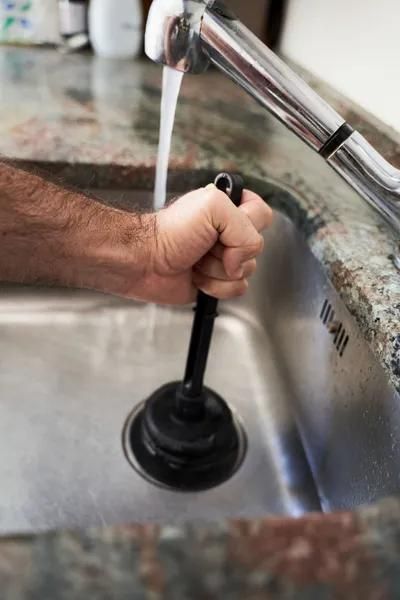 Hand using a plunger in a stainless steel kitchen sink; water running from the faucet.