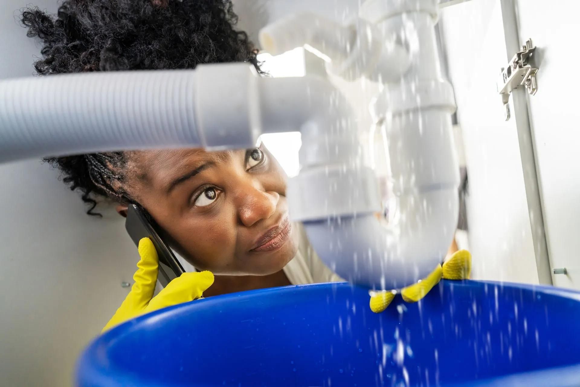 Woman on phone catches water from leaking sink pipe in blue bucket.