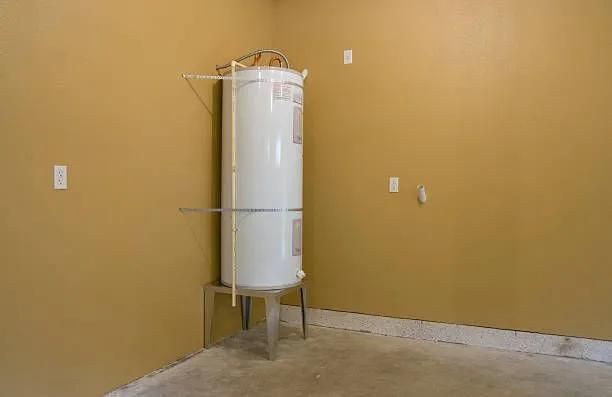 A tall white water heater stands on a metal stand in a corner of a room with tan walls and a concrete floor.