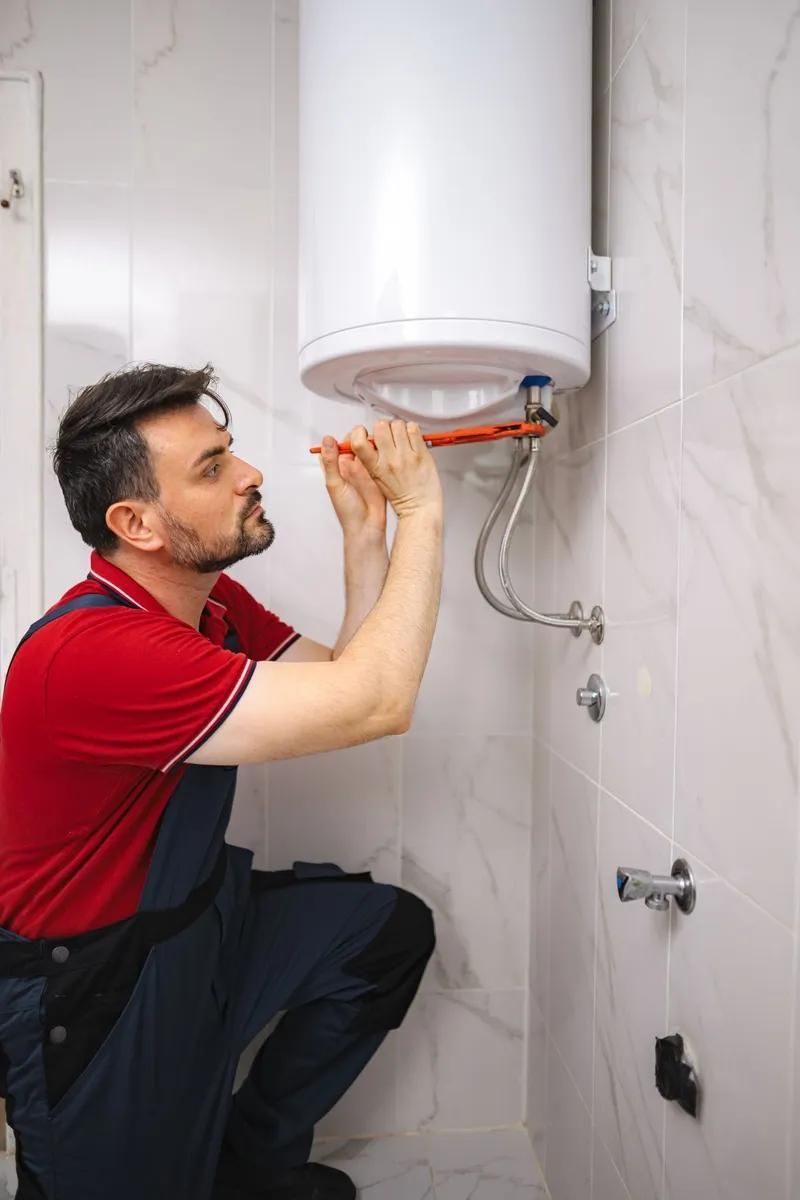 Plumber in red shirt and blue overalls, using a wrench on a water heater in a white-tiled bathroom.