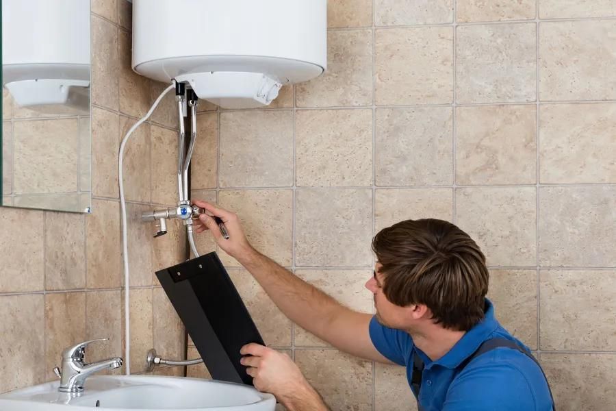 Plumber working on a water heater in a bathroom, holding clipboard, next to a sink.