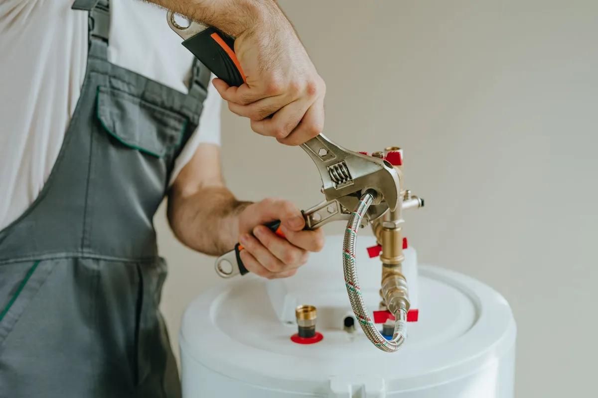 Plumber in gray overalls tightening a fitting on a white water heater with a wrench.