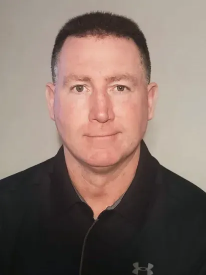 A headshot of a person with short dark hair, wearing a black polo shirt, centered against a plain gray background.