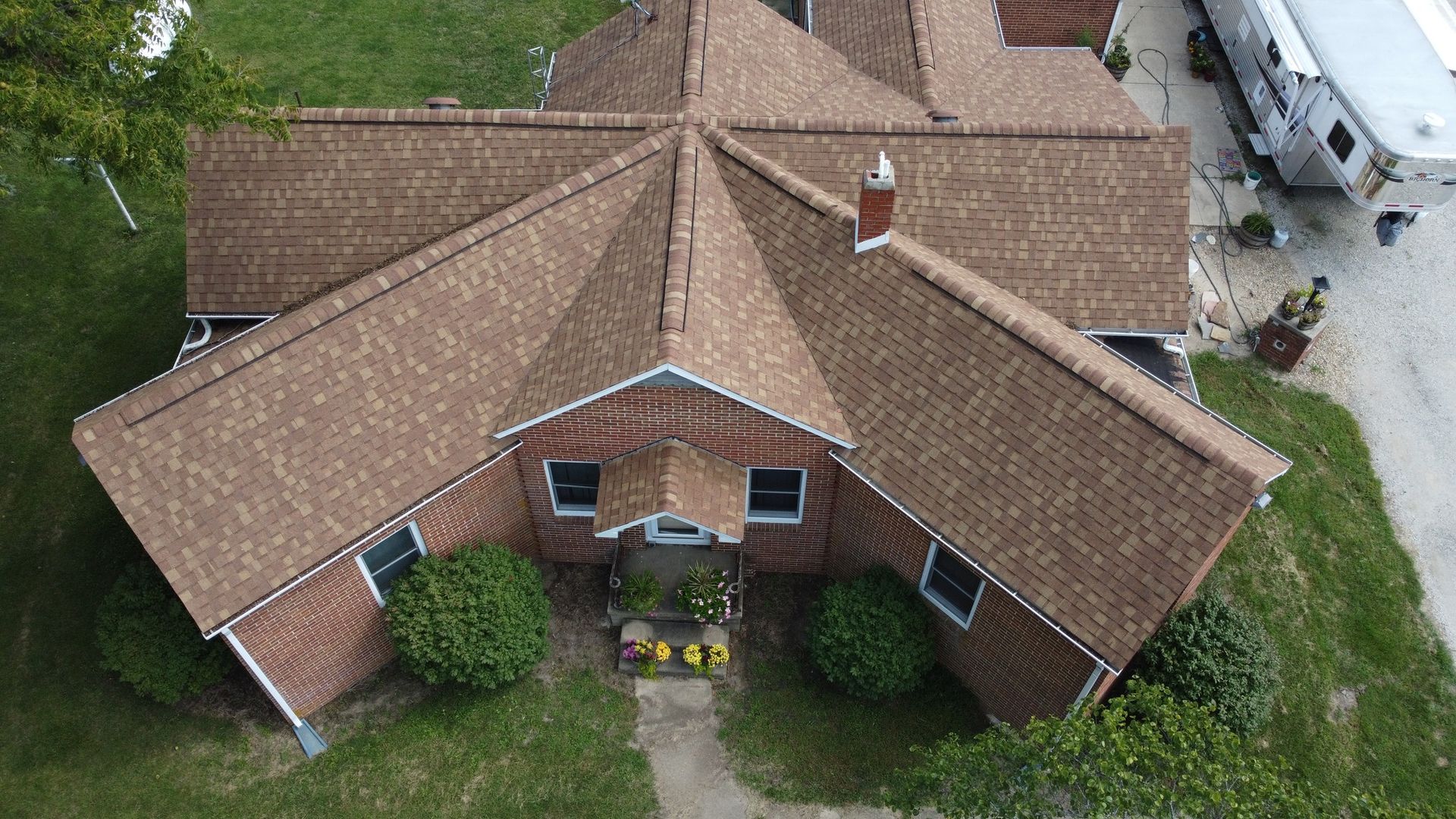 An aerial view of a brick house with a complex, multi-gabled brown shingle roof, surrounded by lawn and green shrubs.
