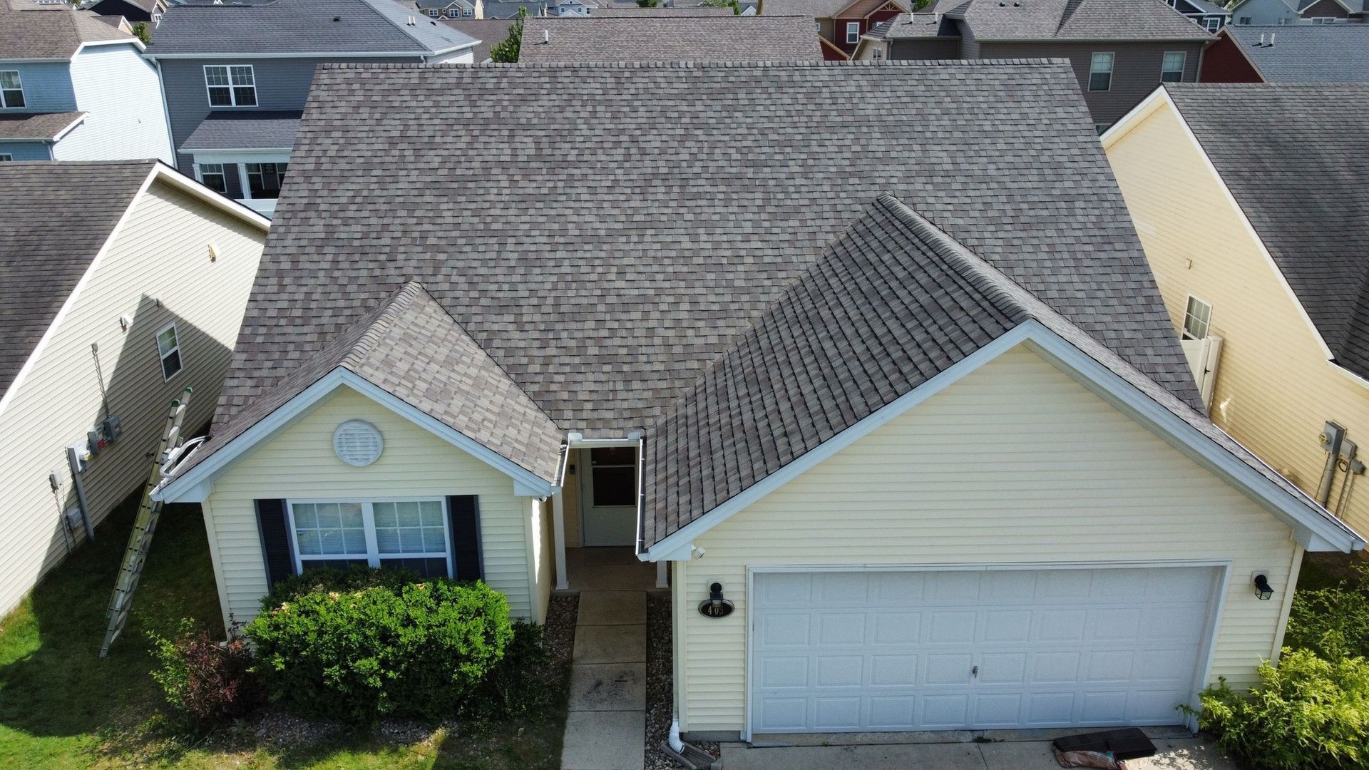An aerial view of a yellow suburban house with a gray shingled roof, a front-facing garage, and a landscaped front yard.