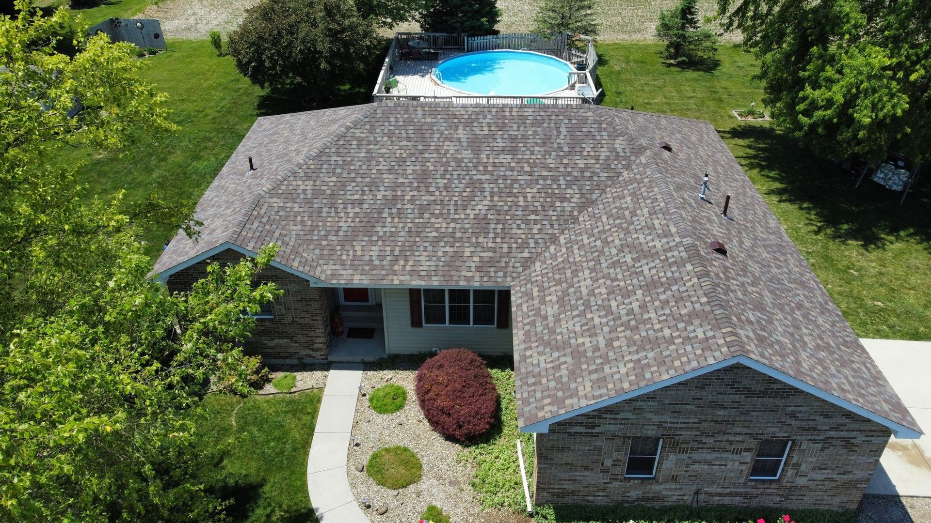 An aerial view of a stone-faced single-story house with a shingled roof, a front lawn, and a backyard swimming pool.
