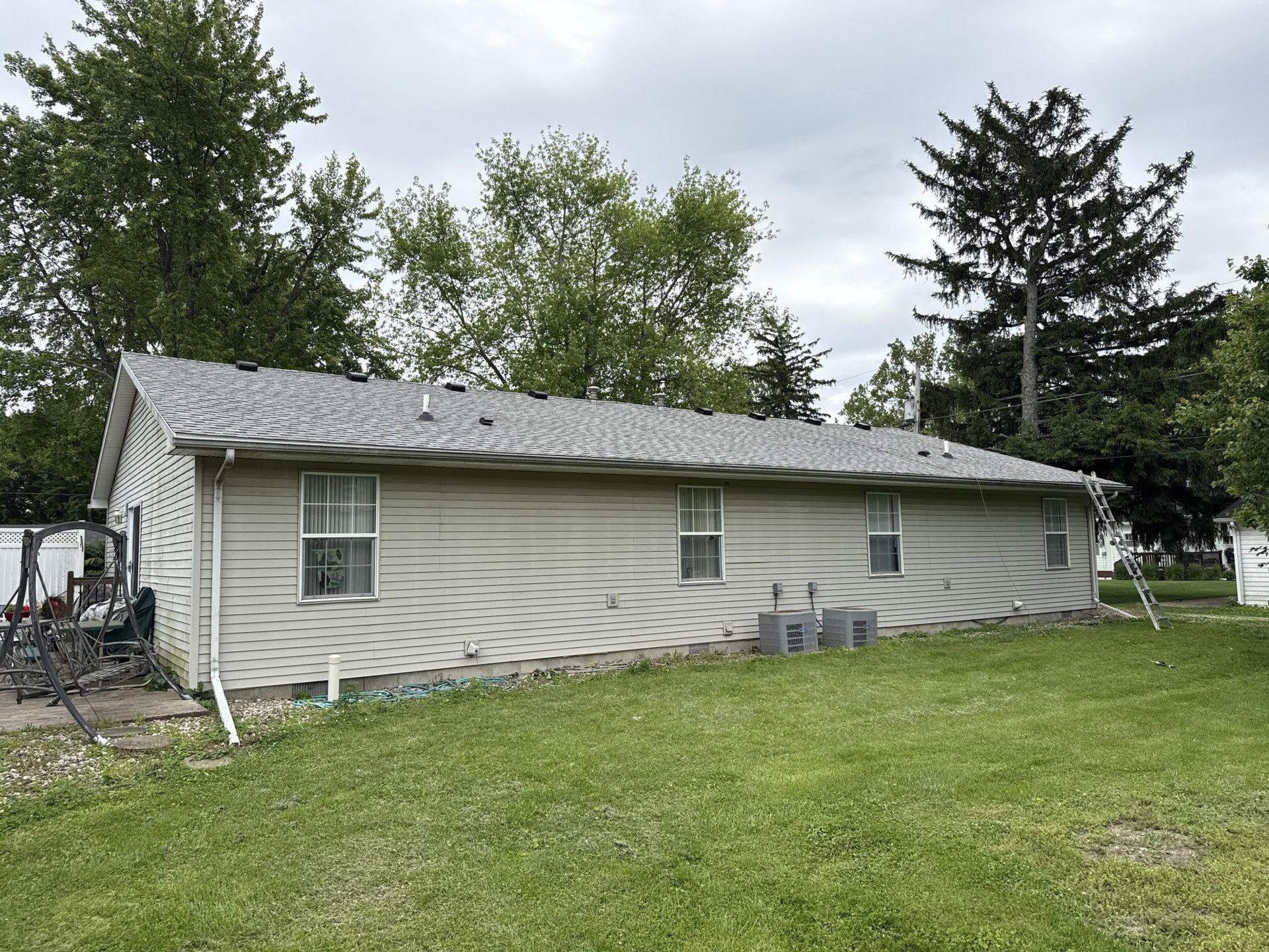 A side view of a beige, single-story house with horizontal vinyl siding and a gray shingled roof, surrounded by green grass.