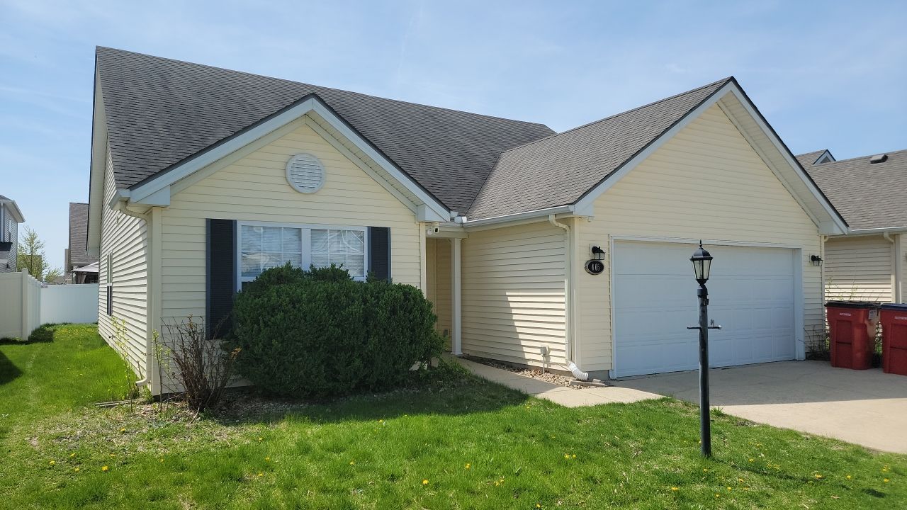A light yellow, single-story suburban house with a gray shingled roof, a two-car garage, and a front lawn.
