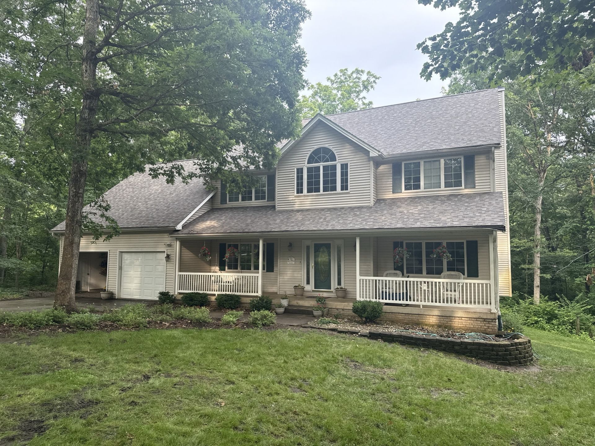 Two-story house with a white front porch and grey shingled roof, surrounded by trees and a green lawn.