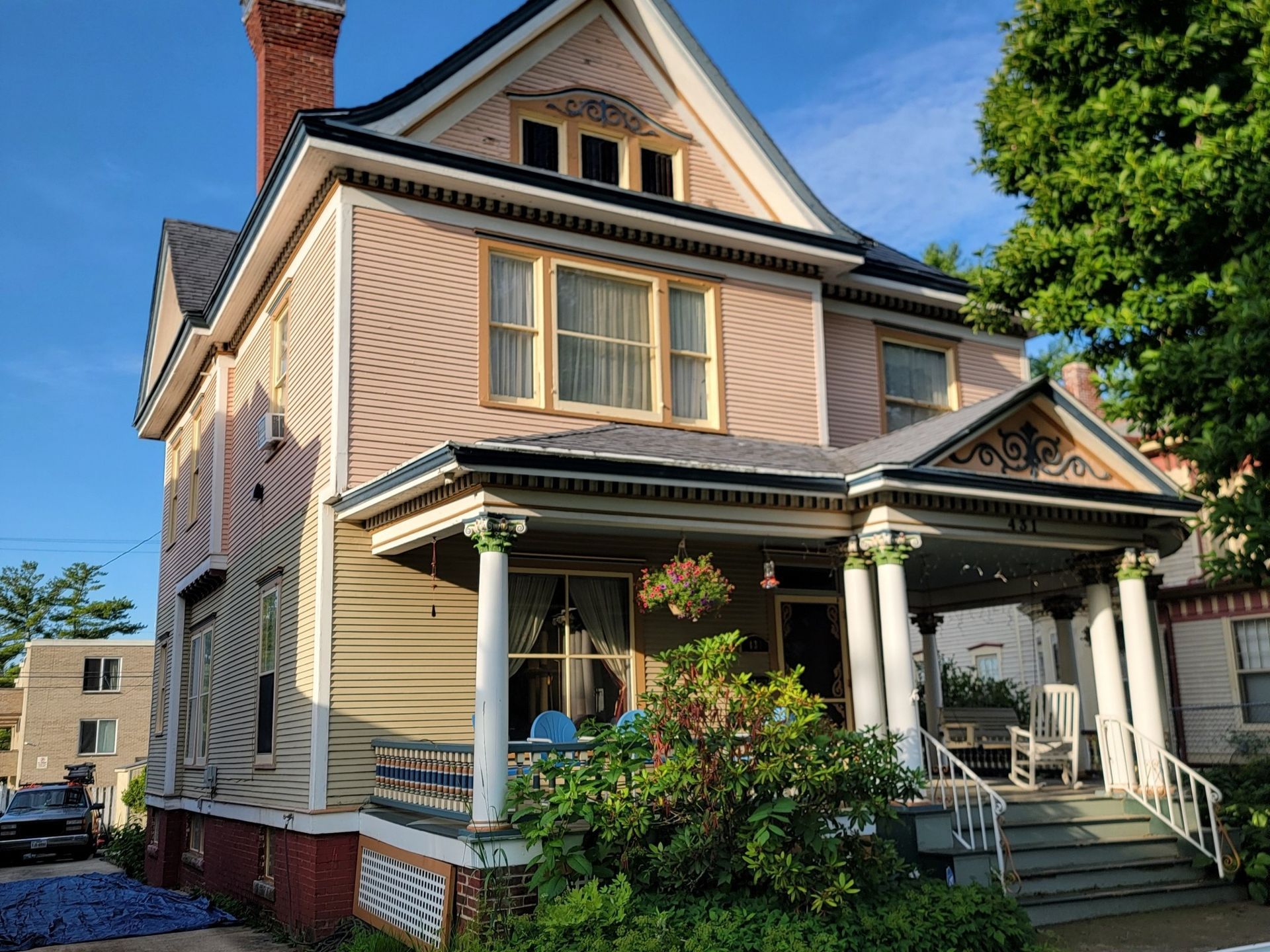 A two-story Victorian house with pale pink siding, tan trim, a large front porch with white columns, and a brick foundation.