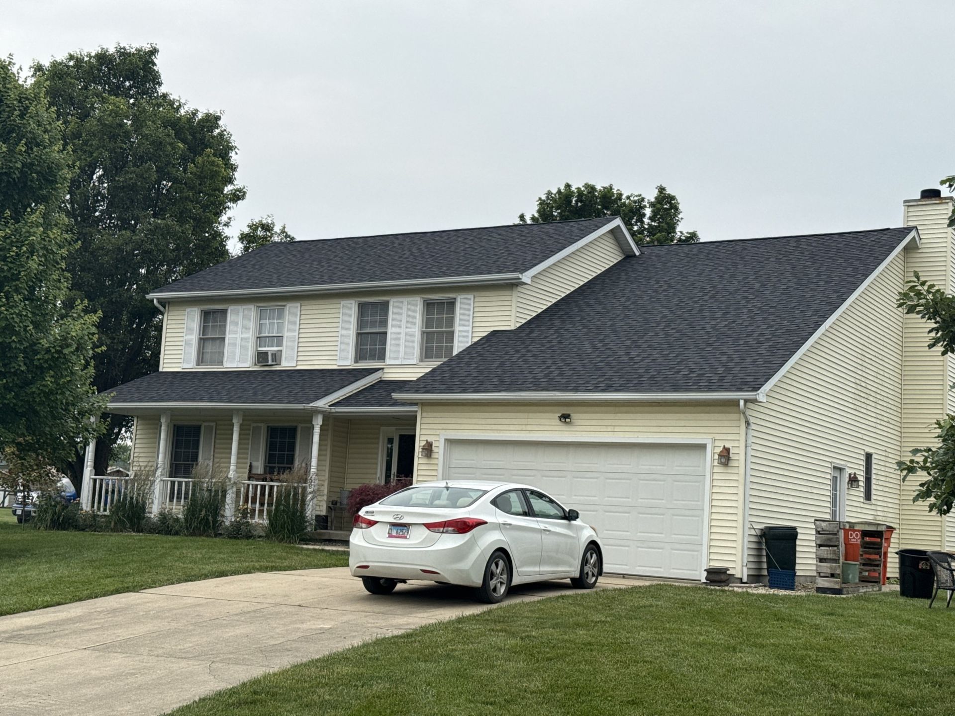 A two-story yellow house with a dark roof and a white garage, featuring a front porch and a white car in the driveway.