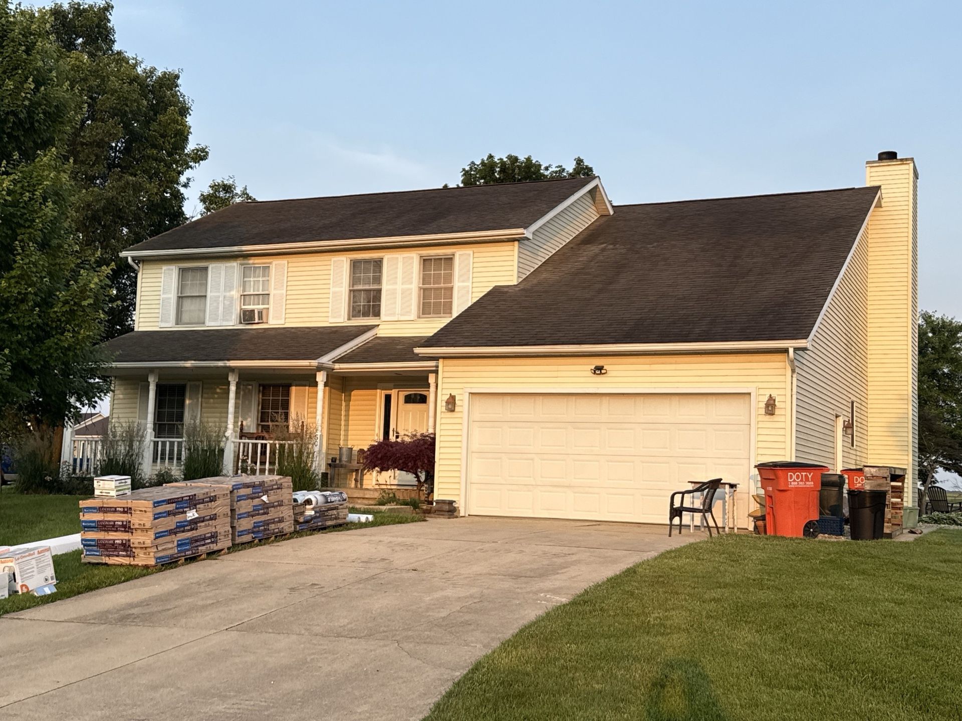 A two-story, yellow house with a white garage and roof-shingle bundles sitting on the driveway.