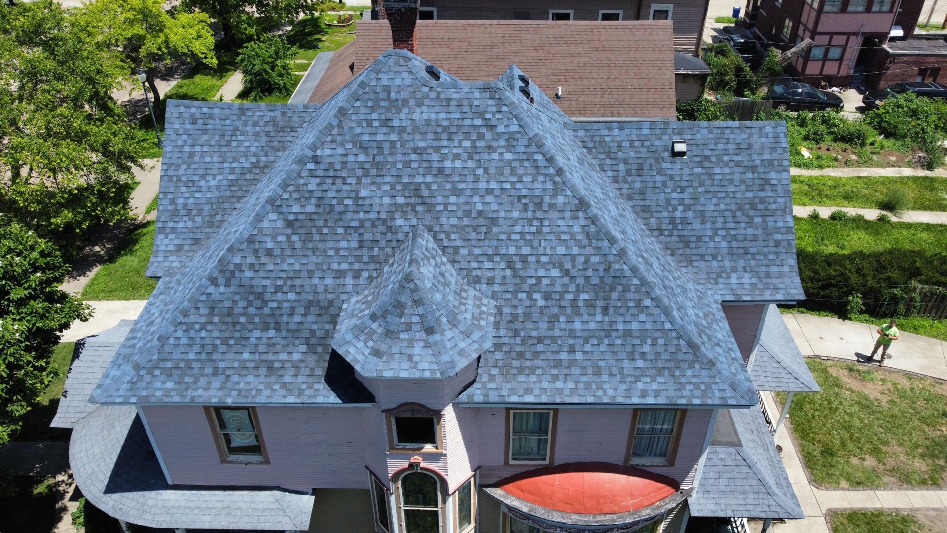 An aerial view of a pink house with a new blue shingled roof, showing complex rooflines and surrounding landscaping.