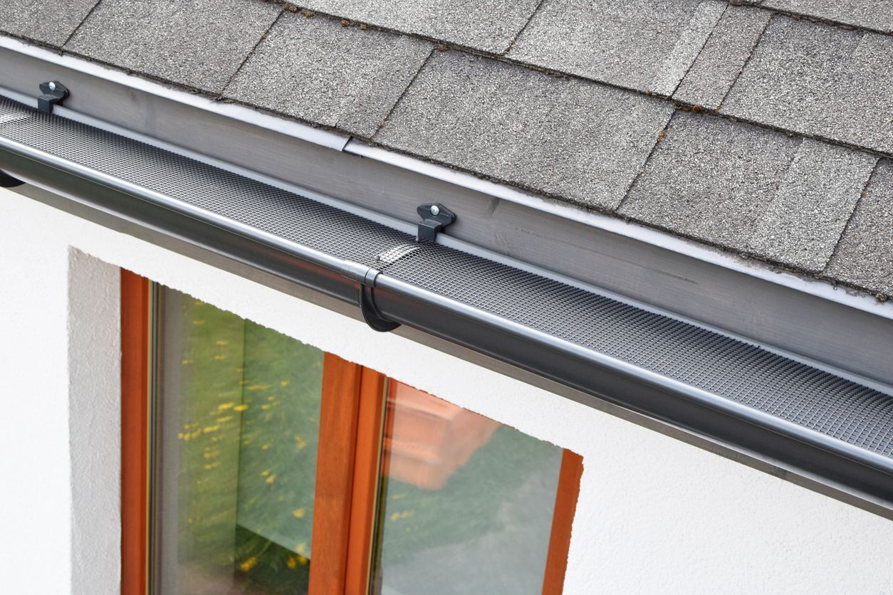 A close-up view of a dark metal gutter covered with a protective mesh screen installed along the edge of a shingled roof.