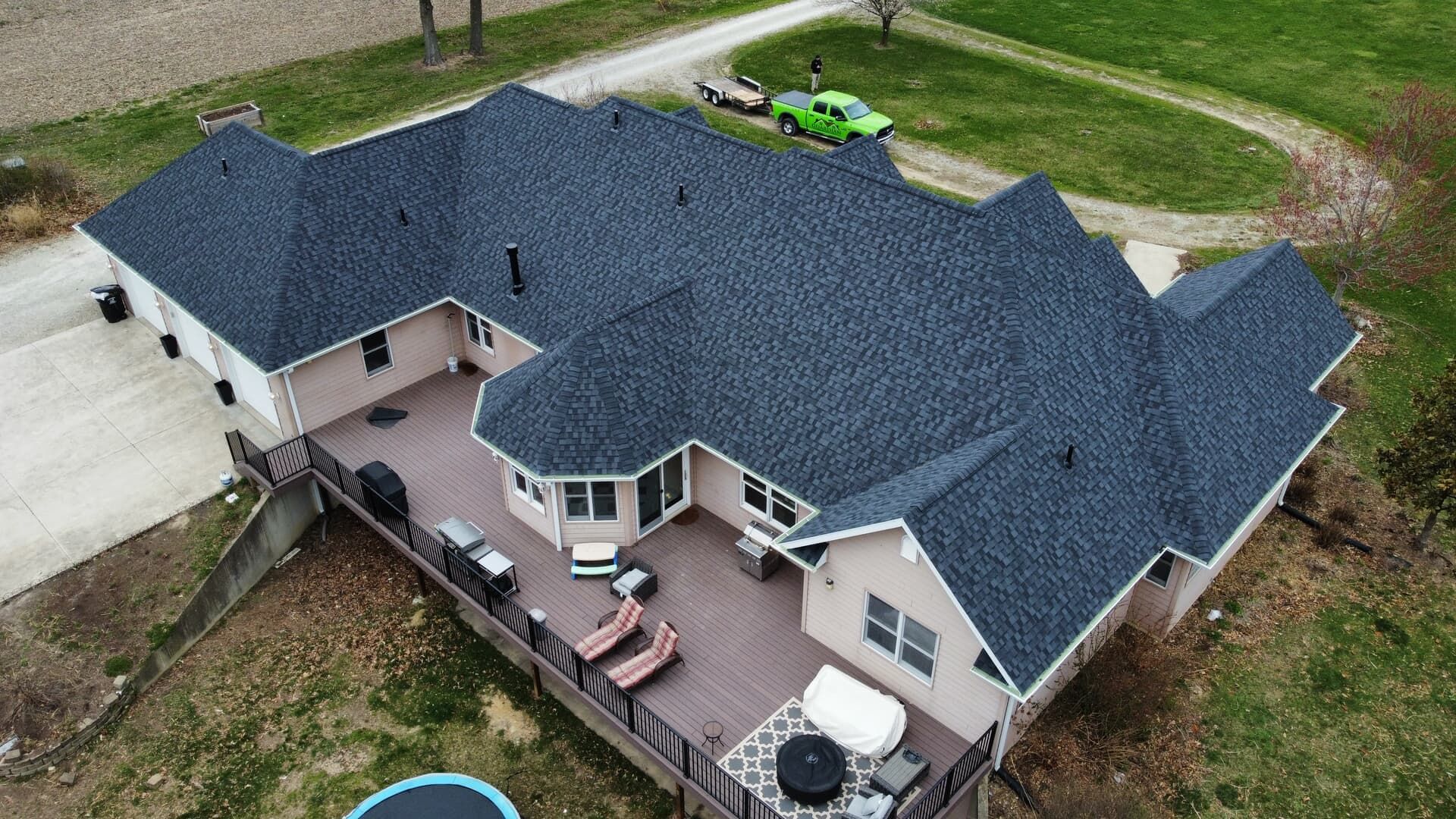 An aerial view of a suburban home with a dark grey roof and a back deck, featuring a hot tub and seating on a lawn.