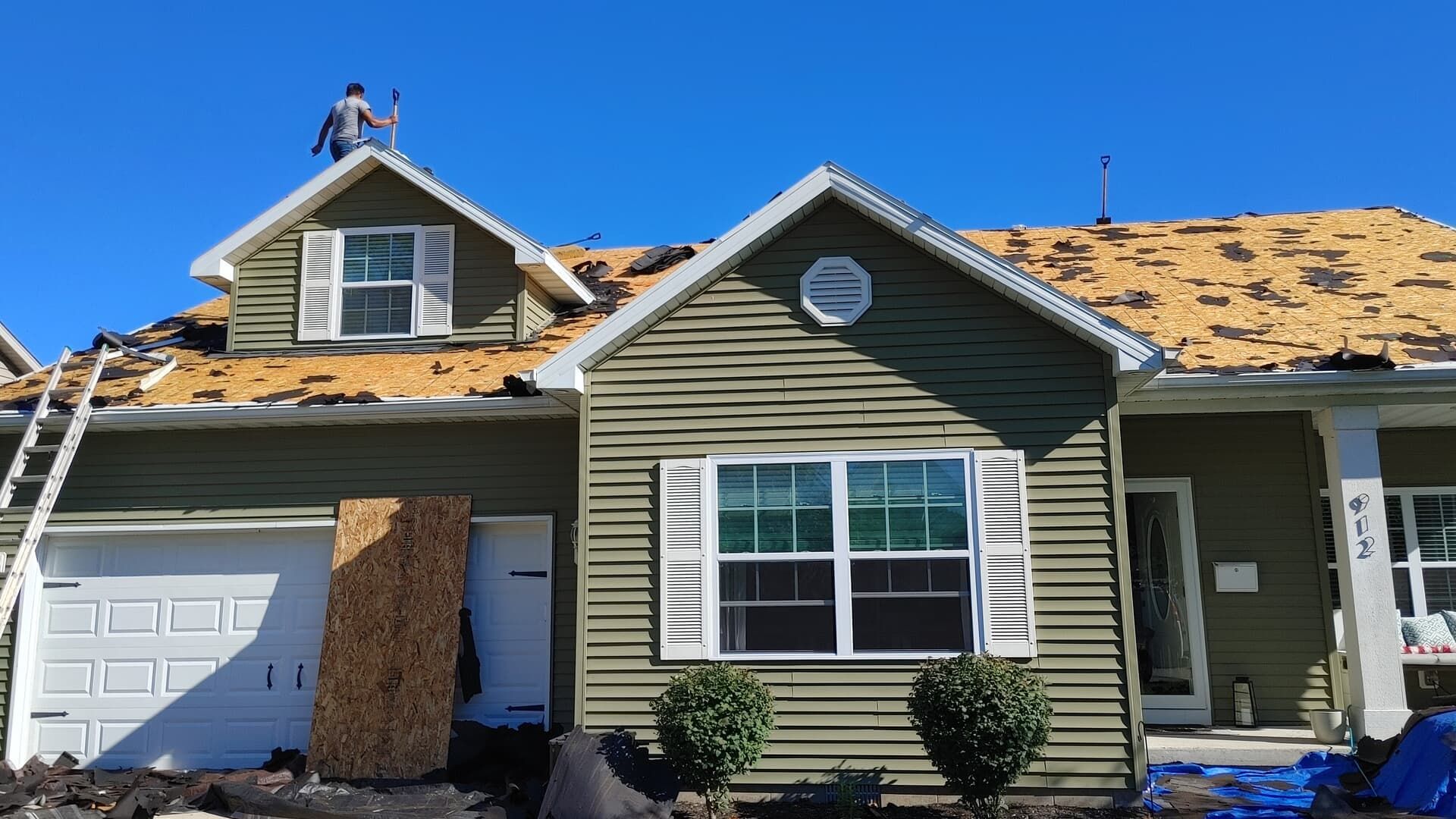 A person works on the roof of a green house with exposed wooden decking while shingles are being replaced.