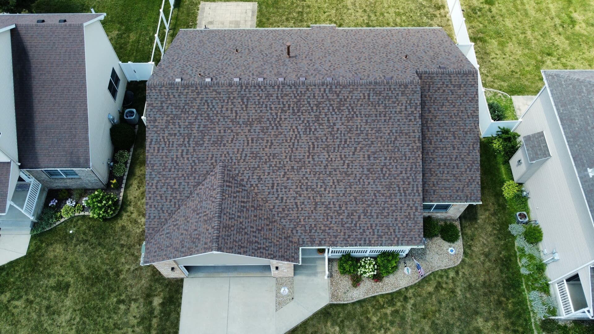 Aerial view of a residential house with a dark brown shingled roof, surrounded by green lawns and white fencing.