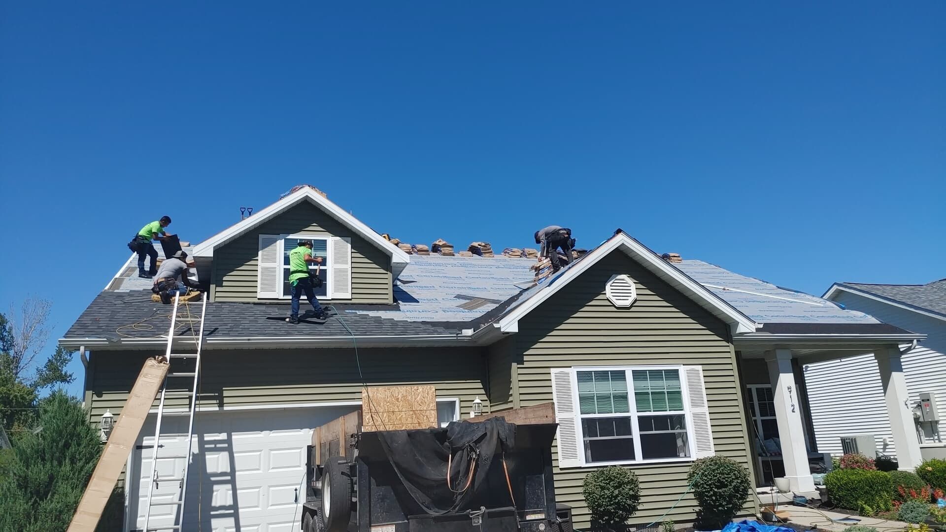 A team of workers in bright safety vests replacing shingles on the roof of a suburban house on a clear, sunny day.
