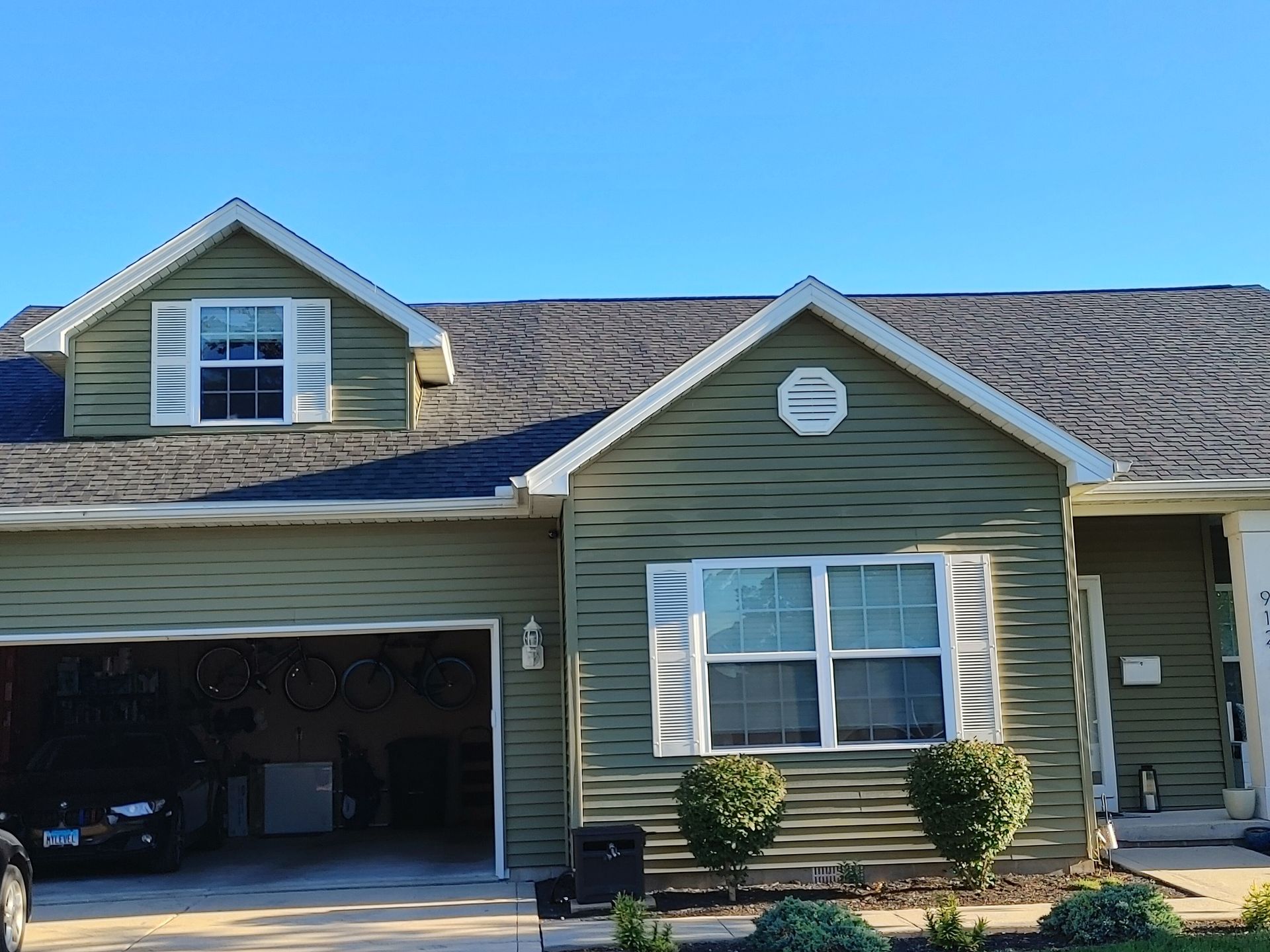 A single-story, olive green vinyl-sided house with a garage, a dormer window, and a gabled front section under a blue sky.