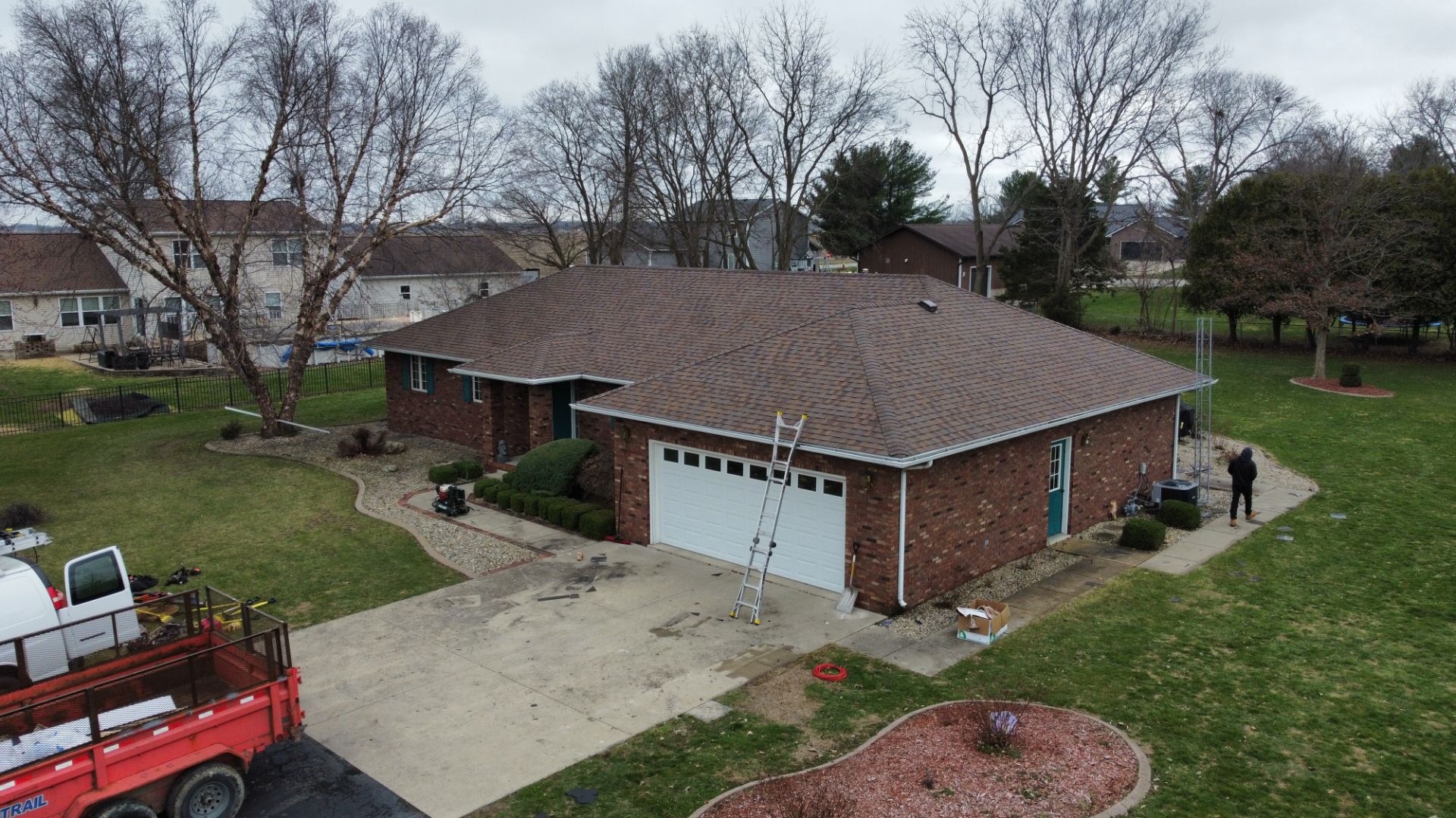 An aerial view of a brick house with a new shingled roof, a white garage door, a ladder, and a truck parked in the driveway.