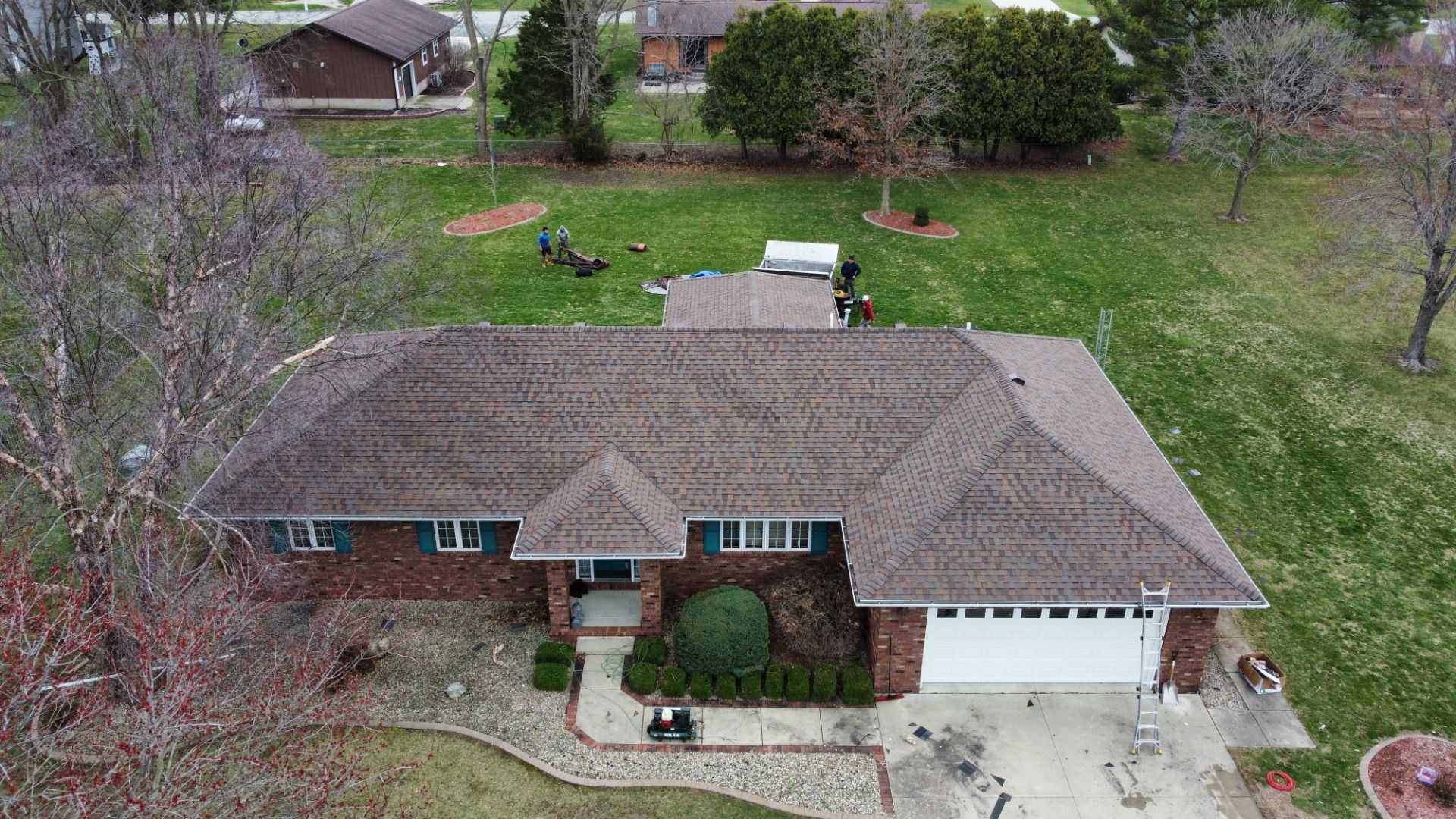 An aerial view of a single-story brick house with a brown shingled roof, a two-car garage, and a large grassy backyard.