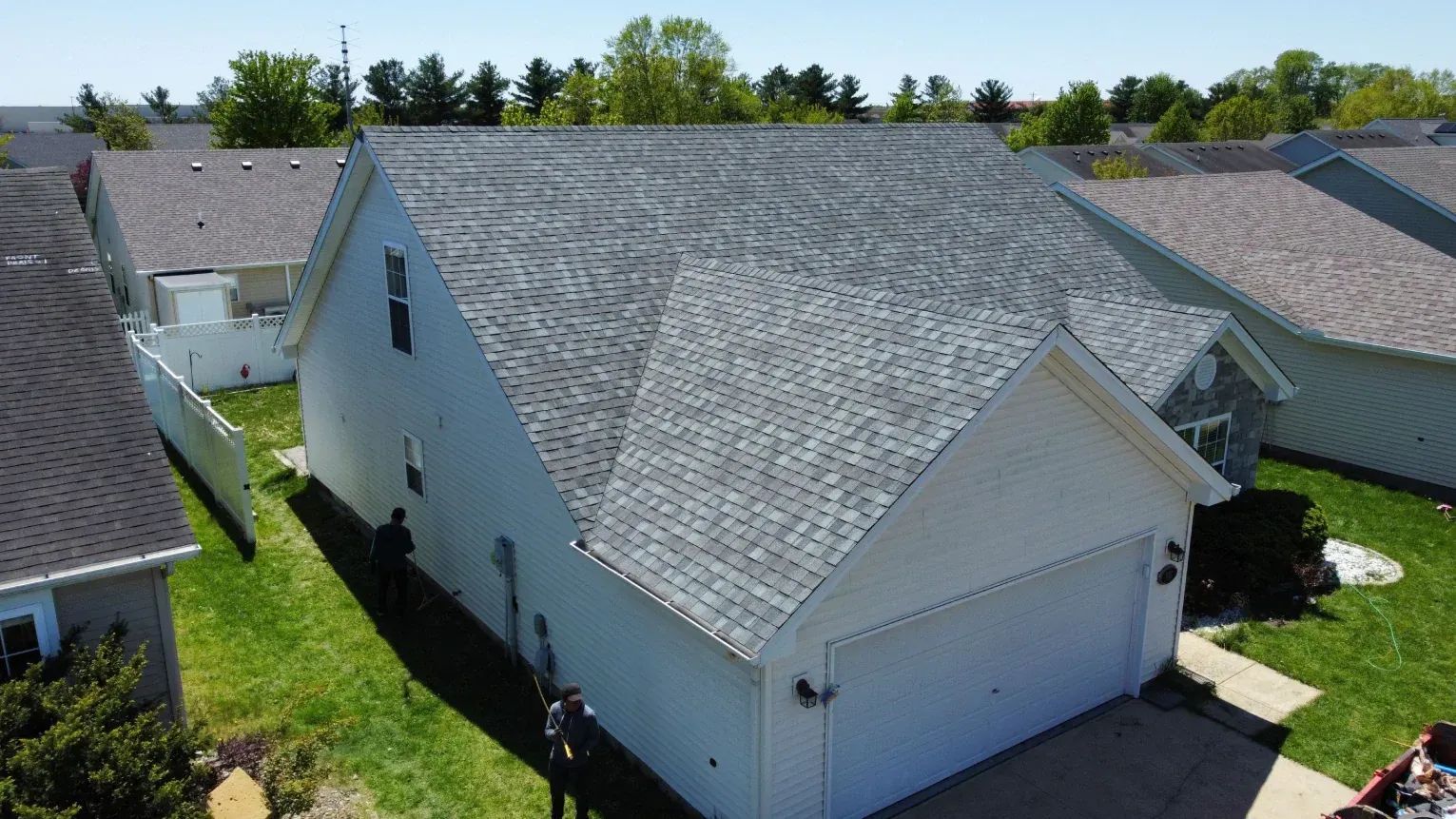 Suburban house with gray shingled roof and white exterior, viewed from above in a sunny neighborhood