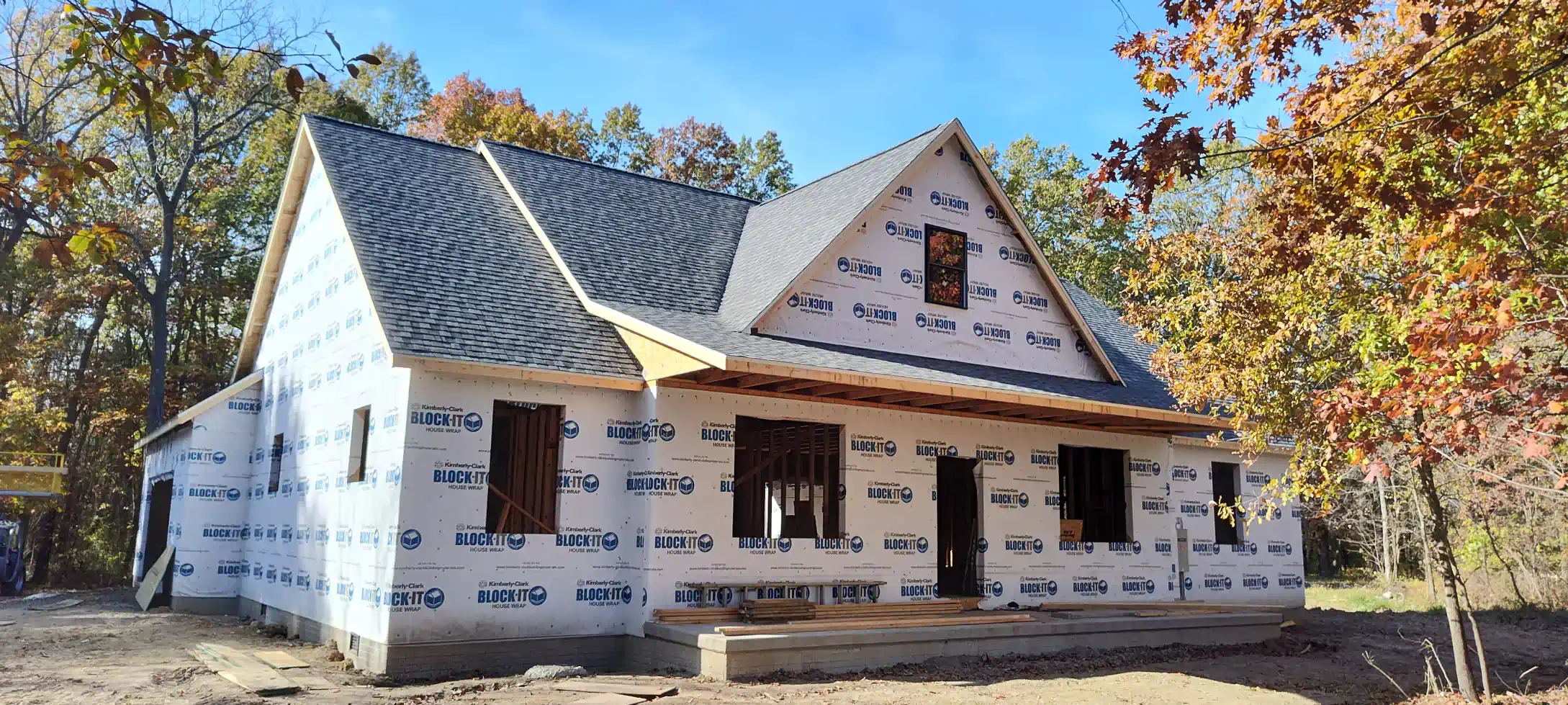A house under construction with a grey shingled roof, white house wrap siding, and exposed window and door frames.